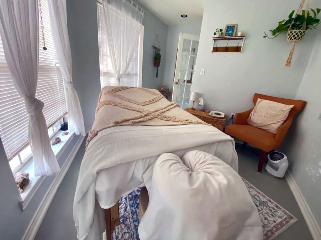 A peaceful, light-filled room set up for massage or wellness treatments. A cushioned table with a cozy beige throw blanket and face cradle is centered in the space, surrounded by soft white curtains, houseplants, and calming decor. To the right, there’s a burnt orange accent chair with a large pillow, a wicker side table with a small lamp, and a wall-mounted shelf. Crystals and decorative items line the window sills, adding to the tranquil, spa-like atmosphere.
