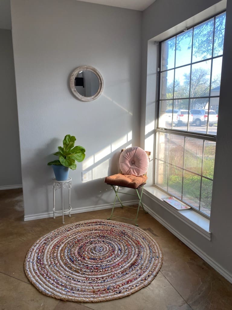 A cozy corner near a large window with natural sunlight streaming in. The space features a round, multicolored braided rug on a polished concrete floor, a light green metal chair with two plush cushions—one peachy-orange and one pink—and a small plant in a white metal stand. A round, rustic-style mirror hangs on the light gray wall, and a magazine or brochure rests on the window sill. Outside the window, cars are parked and trees are visible, adding to the calm, welcoming atmosphere.