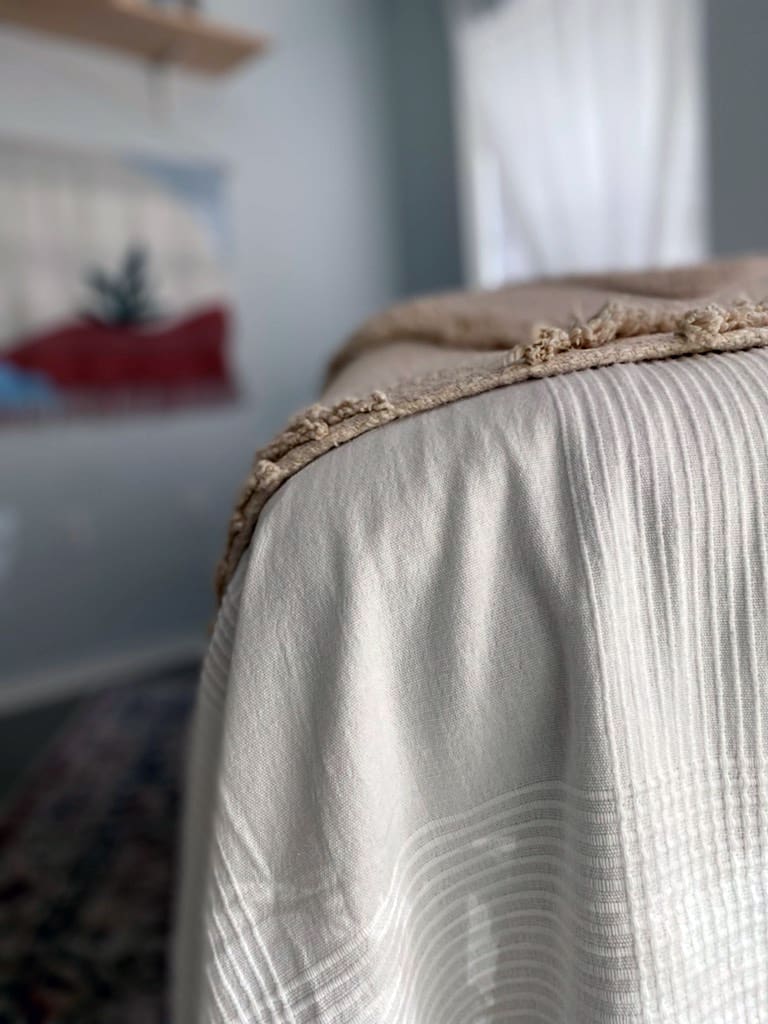 A close-up view of the edge of a massage or treatment table covered with a white textured blanket and a tasseled beige throw. The soft lighting and shallow depth of field create a peaceful, inviting atmosphere. In the blurred background, sheer curtains and a woven wall hanging add to the room's calming aesthetic.