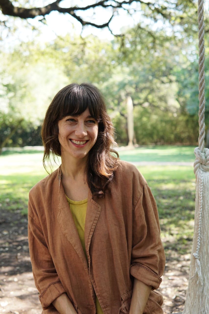 Kendra Bremer, a woman with wavy brown hair and bangs smiles warmly while sitting outdoors. She is wearing a light brown jacket over a yellow top. Sunlight filters through the trees in the background, casting a soft glow on the grassy park setting. A thick rope from a nearby swing is visible on the right side of the image.